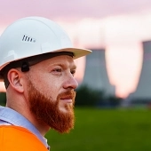 A man with a beard in a protective helmet and an orange vest stands against the background of a nuclear power